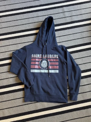 Saint Laurent hoodie - Mörkblå hoodie med huva och dragsko, stor magficka och tryck framtill med texten 'Saint Laurent' samt rödvita ränder och emblem. Mjuk bomullskänsla och avslappnad passform, perfekt för en chill look.