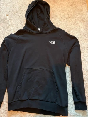 Svart hoodie från The North Face - Svart hoodie från The North Face med vit logga på bröstet och stort tryck på ryggen med texten 'Never Stop Exploring'. Klassisk känguruficka framtill och huva. Tillverkad i mjukt material, perfekt för chill dagar.