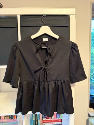 Svart blus med knyt och puffärm - Snygg svart blus med två knyt framtill och lätt puffade ärmar. Blusen har en lös passform och faller fint nedtill. Använd en gång