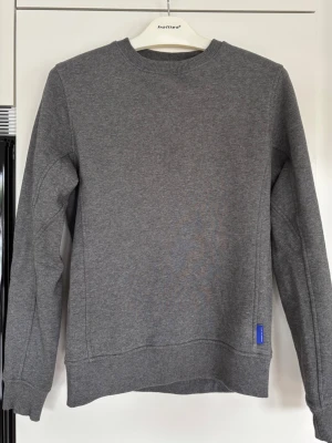 Tiger of Sweden tröja - En stilren grå sweatshirt med rund hals och ribbade muddar vid ärmslut och nederkant. Tröjan har en diskret blå detalj nertill och är tillverkad i mjukt sweatshirtmaterial. Perfekt för en avslappnad look.