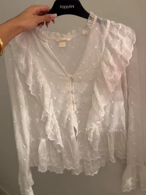 Vit romantisk blus med volanger - Söt vit blus i transparent material med broderade detaljer och volanger längs fram och ärmslut. Blusen har v-ringning, knäppning framtill och långärmad design. Perfekt för en trendig och feminin look.