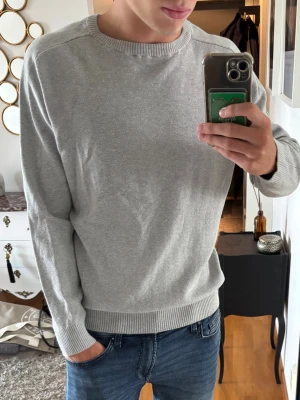 Ljusgrå stickad tröja från Pelle p - En stilren ljusgrå stickad tröja med rund halsringning och ribbade muddar vid ärmslut och nederkant. Perfekt till jeans eller över en skjorta för en clean look. L fits M