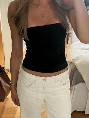 Svart bandeau tubtopp - Säljer en svart bandeau tubtopp som är tight och sitter snyggt på kroppen. Toppen är ärmlös och axelbandslös, perfekt till sommaren. Materialet känns mjukt och stretchigt, troligen bomull eller syntet. Enkel och stilren design som är lätt att matcha.