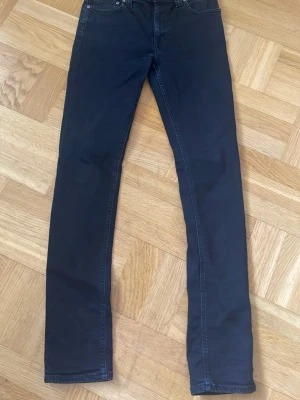 Nudie Jeans, Grim Tim - Säljer ett par svarta slim fit jeans med klassisk femficksmodell och raka ben. Jeansen har en stilren look och är perfekta för dig som gillar en enkel men snygg stil. Materialet känns mjukt och stretchigt för extra komfort.
