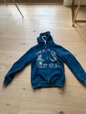 Blå hoodie med tryck och dragkedja - Säljer en blå hoodie med dragkedja och huva. På framsidan finns ett tryck med texten 'Aim for the moon' samt grafik av en tecknad figur och månen. Hoodien har ribbade muddar vid ärmar och nederkant. Perfekt för en chill och avslappnad stil.