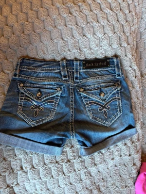 Blå jeansshorts från Rock Revival - Snygga blå jeansshorts från Rock Revival med uppvikta ben och dekorativa bakfickor med broderi och knappar. Shortsen har slitningar, kontrastsömmar och klassisk femficksdesign. Perfekta för en avslappnad och trendig look.