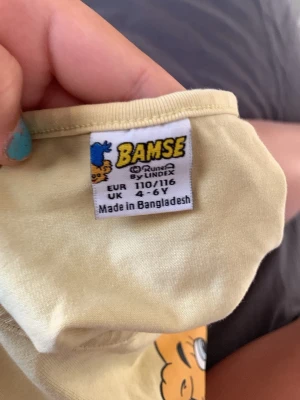 Gul Bamse-klänning från Lindex - Söt gul klänning från Lindex med Bamse-tryck. Klänningen har lång ärm och en mjuk överdel i bomull samt en fluffig kjol i tunt tylltyg. Perfekt för dig som gillar Bamse och vill ha något bekvämt och lekfullt.