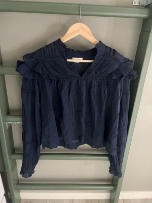 Mörkblå volangblus från Gina Tricot - Supersnygg mörkblå blus från Gina Tricot med volangdetaljer vid axlarna och v-ringning. Blusen har lång ärm och är gjord i ett tunt, krinklat tyg som ger en avslappnad men trendig look. Perfekt att styla med jeans eller kjol.