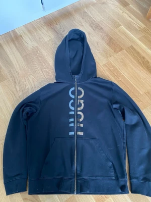 Svart zip-hoodie med HUGO-tryck - Snygg svart hoodie från HUGO med dragkedja framtill och stort vertikalt HUGO-tryck i vitt på framsidan. Hoodien har huva och två fickor framtill. Tillverkad i mjukt bomullsmaterial som känns skönt mot huden.