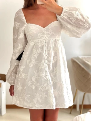 Vit blommig studentklänning med ballongärmar - Söt vit kortklänning med broderade blommor och puffiga långärmar. Klänningen har en v-ringad urringning och markerad midja som ger en feminin siluett. Tillverkad i ett lätt och luftigt tyg, perfekt för varmare dagar. Helt ny med lappar kvar, köpt för 600kr🤍