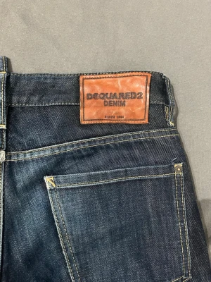 Mörkblå jeans från Dsquared2 - Snygga mörkblå jeans från Dsquared2. Jeansen har en läderpatch med logga bak i midjan och en röd Dsquared2-tag vid gylfen. Tillverkade i slitstark denim, perfekta för en trendig vardagslook.
