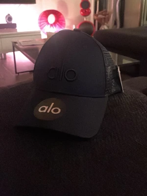Mörkblå keps från Alo - Snygg keps från Alo med broderad logga framtill och mesh på sidorna för extra ventilation. Klassisk böjd skärm och justerbar snapback baktill. Perfekt för en sportig och avslappnad stil.