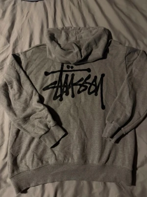 Stussy hoodie - Säljer en grå hoodie från Stüssy med klassisk svart logga på bröstet och stor logga på ryggen. Hoodien har huva med dragsko, magficka och ribbade muddar vid ärmar och nederkant. Perfekt för en avslappnad streetwear-look.