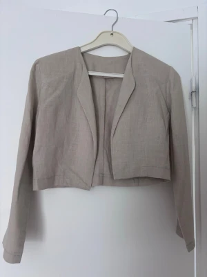 Beige kort kavaj  - Stilren beige kavaj i kort modell med öppen front och långa ärmar. Tillverkad i ett tunt linnetyg som ger en avslappnad och trendig look. Perfekt att matcha med höga byxor eller kjol för en modern outfit.