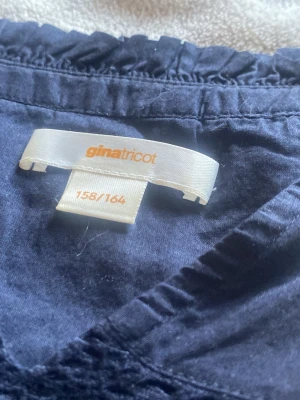Mörkblå blus med volang från Gina Tricot - Snygg mörkblå blus från Gina Tricot med långa ärmar och volangdetaljer på axlarna. Blusen har v-ringning och smockad detalj över bröstet. Tillverkad i ett mjukt och lätt tyg som ger en avslappnad men trendig look.