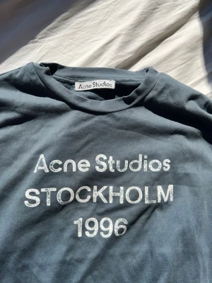 Mörkblå långärmad topp Acne Studios - Snygg mörkblå långärmad topp från Acne Studios med vit text 'Acne Studios STOCKHOLM 1996' framtill. Klassisk rund halsringning och rak passform. Perfekt för dig som gillar stilrena och trendiga plagg.