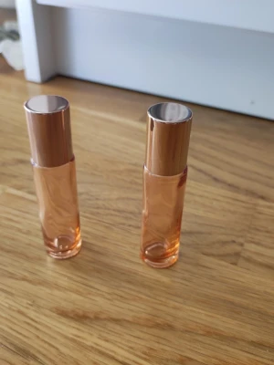 Två små tomma parfymflaskor med roséguldlock - Säljer två stilrena tomma parfymflaskor i genomskinligt orange glas med eleganta roséguldfärgade lock. Flaskorna har rund form och roll-on applikator, perfekta att ha i väskan. Passar dig som gillar smidiga och snygga parfymförpackningar.