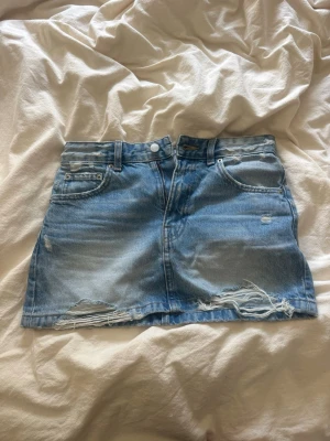Blå jeanskjol med slitningar - Säljer en kort blå jeanskjol med slitna detaljer och fransar längst ner. Kjolen har klassiska fem fickor, bälteshällor och dragkedja med knapp framtill. Perfekt för dig som gillar en avslappnad och trendig stil.
