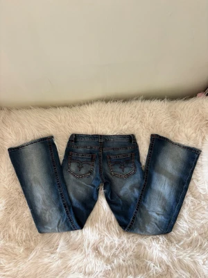 Blå bootcut jeans med broderade fickor - Säljer ett par blå bootcut jeans med snygg slitning och broderade detaljer på bakfickorna. Jeansen har klassisk femficksmodell.