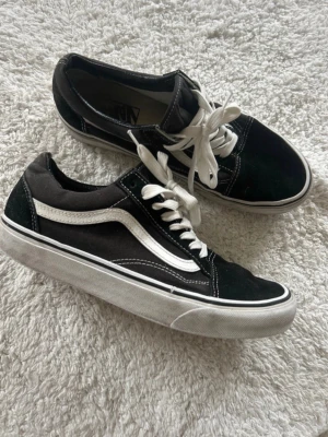 Svarta Vans Old Skool sneakers - Klassiska Vans Old Skool sneakers i svart mocka och canvas med vit sula och vit sidostripes. Skorna har vita snören och kontrastsömmar som ger en snygg retrolook. Perfekta för dig som gillar streetwear och skate-stil.