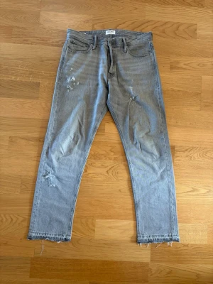 Gråa slitna raka jeans.  - Säljer ett par ljusblå jeans med raka ben och slitningar på både fram- och baksida. Jeansen har fransiga benslut och klassisk femficksmodell. Materialet är denim i bomull och de har en avslappnad vibe som passar perfekt till streetwear. Storlek: 32/32