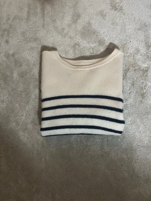Vit stickad tröja med marinblå ränder - Klassisk vit stickad tröja med marinblå horisontella ränder. Tröjan har rund halsringning och långa ärmar. Perfekt för dig som gillar en tidlös och stilren look. Materialet ser mjukt och bekvämt ut, passar till många olika outfits.