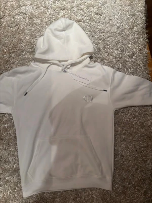 Di0r hoodie vit - Snygg vit hoodie med klassisk huva och dragsko. Framtill finns en diskret CD-logga broderad på bröstet och en stor magficka. Hoodien har ribbade muddar och är tillverkad i mjukt material för maximal komfort.