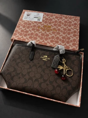 Brun monogram clutch - Snygg brun clutch med klassiskt monogrammönster och guldfärgad logga. Väskan har svarta handtag och en dekorativ nyckelring med körsbärsdetaljer i rött och guld. Perfekt accessoar för dig som gillar stilrena detaljer.