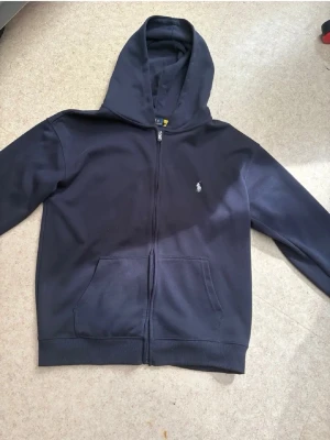 Mörkblå hoodie från Polo Ralph Lauren - Snygg mörkblå hoodie från Polo Ralph Lauren med dragkedja framtill och klassisk logga på bröstet. Hoodien har känguruficka och ribbade muddar. Perfekt för en chill och stilren look. Materialet känns mjukt och bekvämt.