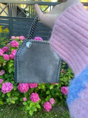 Grå Stella McCartney handväska - Säljer en snygg grå handväska från Stella McCartney med kedjedetaljer och mjukt mockaliknande material. Väskan har en silverfärgad kedja som axelrem och dekorativ kant, samt en rund logotypbricka. Perfekt storlek för det viktigaste.
