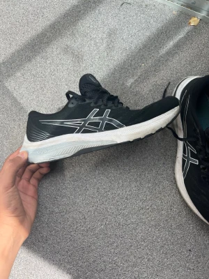 Svarta Asics sneakers med vit sula - Svarta sneakers från Asics med vit sula och klassisk logga i vitt på sidan. Skorna har mesh-ovandel för bra andningsförmåga och snörning framtill. Perfekta för dig som gillar sportig stil och vill ha bekväma skor till vardags.