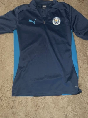 Manchester City träningströja Puma - Mörkblå Manchester City träningströja från Puma med ljusblå detaljer på sidorna. Halvlång dragkedja framtill och klubbmärke på bröstet. Tillverkad i funktionsmaterial som andas, perfekt för träning eller match.