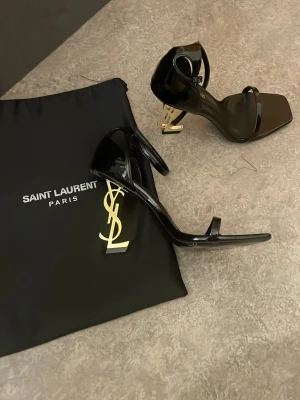 Svarta Saint Laurent sandaler med YSL-klack - Säljer ett par svarta sandaler från Saint Laurent med ikonisk YSL-logga i guld på klacken. Sandalerna har smala remmar, fyrkantig tå och är tillverkade i blankt läder. Perfekta för dig som vill sticka ut med en lyxig och trendig look.
