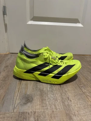 Adidas Adizero Pro 4 löparskor neongula (Kolfiber) - Säljer ett par neongula Adidas Adizero löparskor med svarta ränder på sidorna och grå detaljer vid hälen. Skorna har lättviktssula med texten 'Lightstrike Pro' och slitstark yttersula. Perfekta för dig som vill springa snabbt och sticka ut.