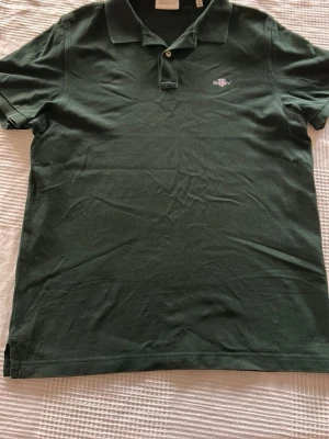 Mörkgrön pikétröja från GANT - Snygg mörkgrön pikétröja från GANT i regular fit. Klassisk krage, kort ärm och två knappar framtill. Liten broderad logga på bröstet. Tillverkad i mjuk bomull som känns skön mot huden.