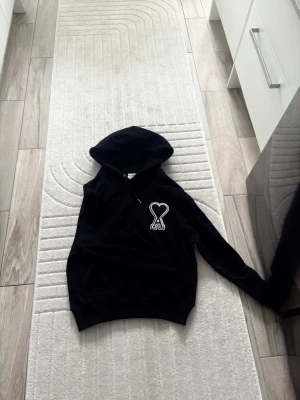 Ami hoodie - Svart hoodie med huva och dragsko. På bröstet finns en vit hjärtformad logga med bokstaven A. Hoodien har långa ärmar och ribbade muddar vid ärmslut och nederkant. Perfekt för en avslappnad och trendig stil. Det sär storlek S men sitter som M.