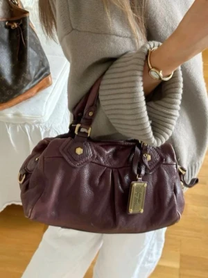 Vinröd handväska Marc by Marc Jacobs - Säljer en vinröd handväska i mjukt läder från Marc by Marc Jacobs. Väskan har guldfärgade metalldetaljer, dubbla handtag och avtagbar axelrem. Klassisk design med dekorativa sömmar och logoplatta framtill. Perfekt för dig som vill ha en snygg och rymlig väska.