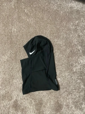 Riktig Nike ski mask - Säljer en svart balaclava från Nike Pro med vit logga på sidan. Tillverkad i ett stretchigt och lätt material som andas, perfekt för träning eller kalla dagar. Snygg och sportig design med synliga sömmar och diskret etikett.