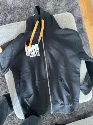 Svart MM Sports hoodie med dragkedja - Svart hoodie från MM Sports med vit logga på bröstet och ärmen. Hoodien har orangea snören i huvan, dragkedja framtill och två fickor. Tillverkad i mjukt bomullsmaterial, perfekt för en sportig och avslappnad stil.