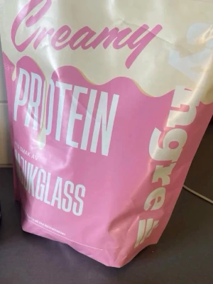 Creamy proteinpulver mjukglassmak - Stor påse proteinpulver med smak av mjukglass. Öppnad och tagen två skopor Förpackningen är rosa och vit med text i stora bokstäver. Perfekt för dig som vill boosta träningen med extra protein och gillar söta smaker.