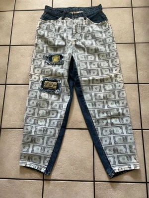 Jeans med dollartryck och patchar storlek 32 - Unika jeans med dollartryck över hela byxorna och kontrasterande mörkblå denim längs insidan av benen. Två slitna patchar med text och denimdetaljer på vänster ben ger extra edge. Perfekta för dig som vill sticka ut och gillar streetwear.