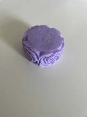 Ljuslila rosformad tvål - Säljer en handgjord tvål i ljuslila färg med vackra rosdetaljer runt sidorna. Tvålen är rund och har en dekorativ design som passar perfekt som present eller dekoration i badrummet. Doftar ljuvligt av syren