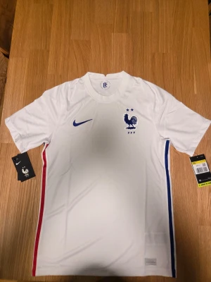 Nike Frankrike matchtröja 2020, storlek S - Vit matchtröja från Frankrike med den franska tuppen och två stjärnor ovanför. Tröjan har en blå Nike-logga på bröstet och en röd och blå rand längs sidorna.