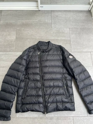 Svart dunjacka från Moncler - Snygg svart dunjacka från Moncler med quiltad design och asymmetrisk dragkedja. Jackan har ståkrage med tryckknapp och två sidofickor med dragkedja. Moncler-logga på vänster ärm ger en exklusiv touch. Perfekt för hösten! Den är knappt använd och i mycket bra skick.