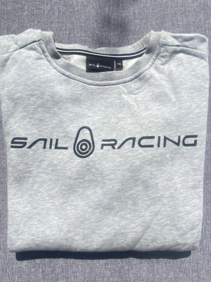 Ljusgrå sweatshirt från Sail Racing strl 160 - Ljusgrå sweatshirt från Sail Racing med rund halsringning och stor svart logotyp på bröstet. Tröjan är tillverkad i mjukt bomullsmaterial och har en klassisk, avslappnad passform. Perfekt för en sportig och stilren look. Köpt på KidsBrandstore