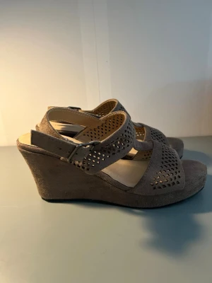 Beige kilklackade sandaler med remmar - Snygga sandaler i beige mockaimitation med kilklack och justerbar rem runt ankeln. De har breda remmar med dekorativa hål och öppen tå. 