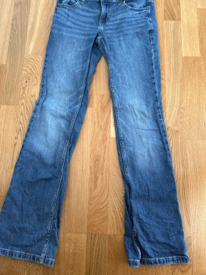Blå bootcut jeans från Lee - Säljer ett par klassiska blå bootcut jeans från Lee med fem fickor och normal midja. Jeansen har snygga slitningar och kontrastsömmar. Perfekta för dig som gillar en avslappnad men trendig look.