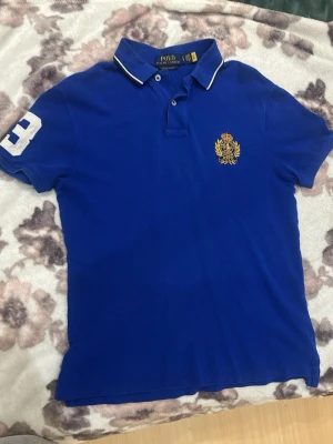 POLO RALPH LAUREN - Klassisk blå pikétröja från Polo Ralph Lauren med vit kant på kragen, broderad logga på bröstet och stort tryck på ryggen. Kortärmad modell med nummerdetalj på ärmen. Perfekt för en sportig och stilren look. HELT ÄKTA, OANVÄND, FICK I PRESENT ETT TAG SEDAN MEN VAR FÖR LITEN. KÖPTES I OFFICIELLA BUTIKEN FÖR 2000 NÅGOT. PASSAR S OCH M