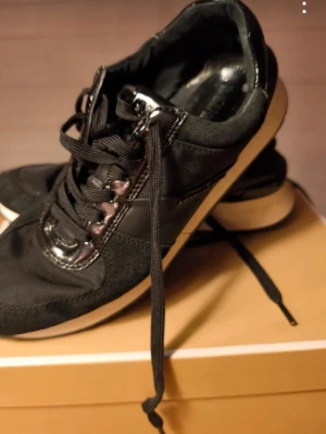 Svarta sneakers med vita sulor - Snygga svarta sneakers med vita sulor och svarta skosnören. Ovandelen är i en mix av textil, mocka och glansiga detaljer som ger en cool kontrast. Klassisk låg modell som passar till det mesta. Skorna är väl använda men sköna sulan i båda är lite skadade se bild.