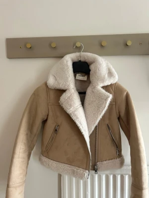 Beige teddyjacka med vit fuskpäls - Snygg beige teddyjacka med vit fuskpäls på krage, ärmslut och nederkant. Jackan har dragkedja framtill och två snedställda fickor med dragkedja. Perfekt för kalla dagar och ger en trendig look.